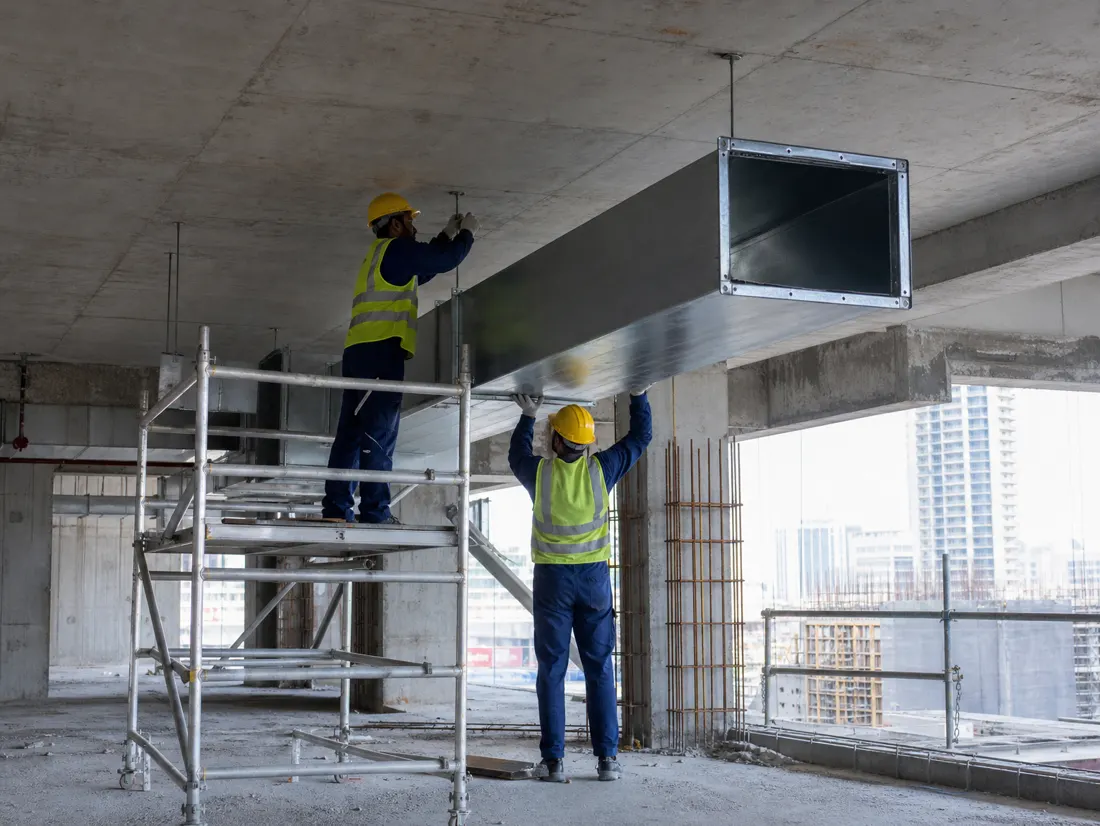 Two technicians installing a galvanised steel duct section into a concrete soffit